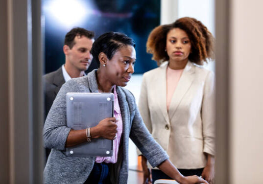 A multi-ethnic group of three business people after a meeting in an office board room. An African-American woman is leaving the room carrying her laptop. Another woman and a man are standing in the background.